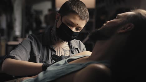 mujer caucásica con máscara facial tatuando los brazos de su cliente.