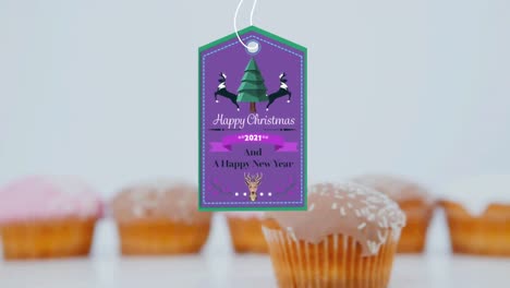 Animation-Von-Weihnachtsgrüßen-Auf-Einem-Etikett-über-Cupcakes-Auf-Weißem-Hintergrund