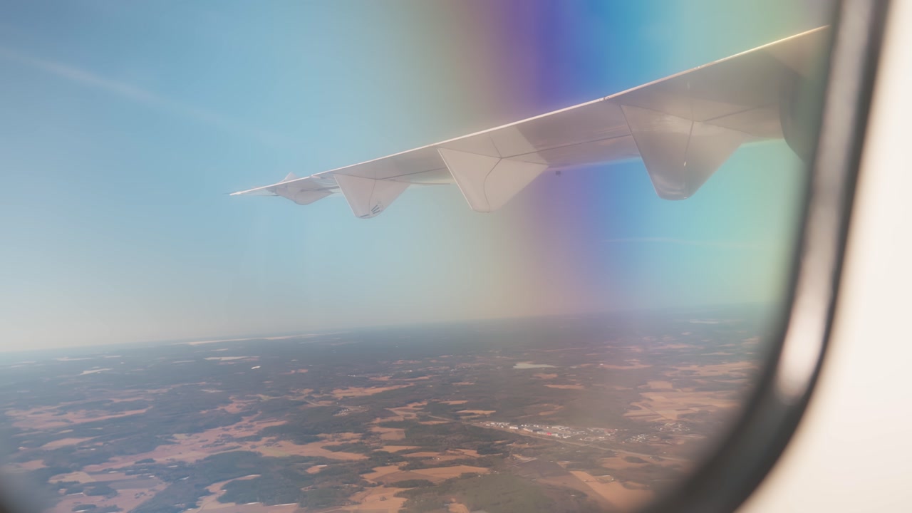 Handheld airplane-window view with wing silhouette and rainbow lens flare, capturing real passenger flight atmosphere above hazy countryside.