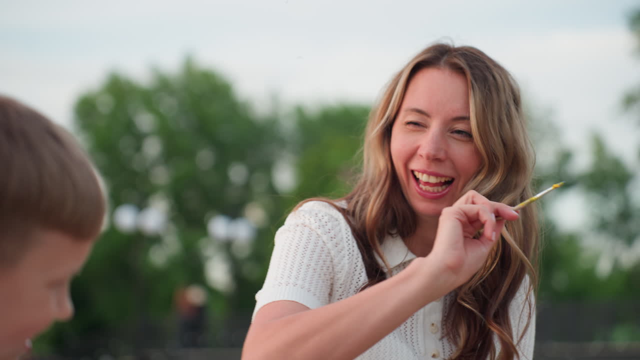 cheerful mom and son seated on wooden dock by pool under sunset glow painting on boards, mother playfully touches son cheek with brush as boy smiles and leans away in warm evening art session
