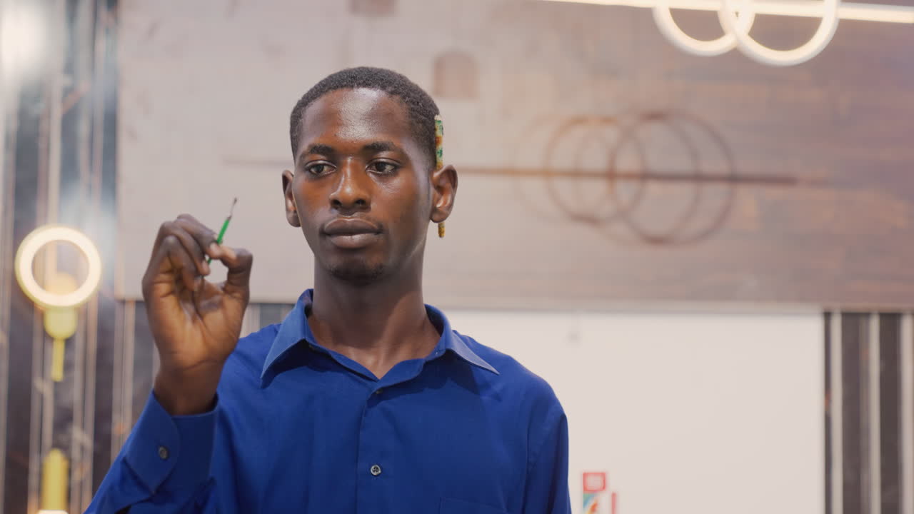 Uomo di colore che dipinge su vetro, artista contemplativo in uno studio moderno, lampade circolari e sfondo con pannelli di legno, primo piano della mano che tiene un pennarello verde, tratti misurati, sguardo pensieroso, pianificazione creativa.