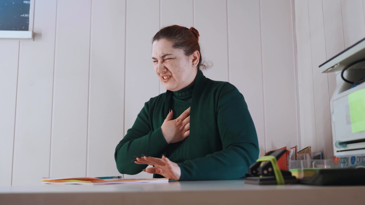 White office manager calling ambulance while writing forms, desk organizer and printer visible, hand on chest between notes, composed yet concerned, emergency protocol in progress