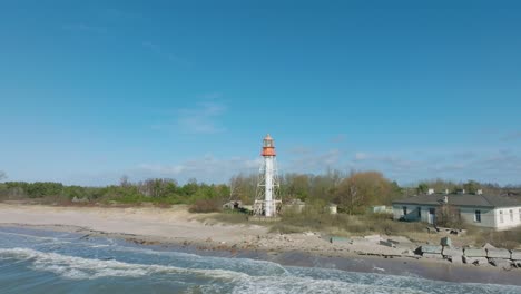 Aerial-establishing-view-of-white-colored-Pape-lighthouse,-Baltic-sea-coastline,-Latvia,-white-sand-beach,-large-waves-crashing,-sunny-day-with-clouds,-wide-ascending-drone-orbit-shot