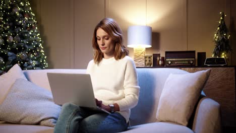 feliz joven hermosa mujer sentada en un sofá en una casa decorada cerca de un brillante árbol de navidad por la noche y escribiendo y navegando en una laptop en la víspera de navidad
