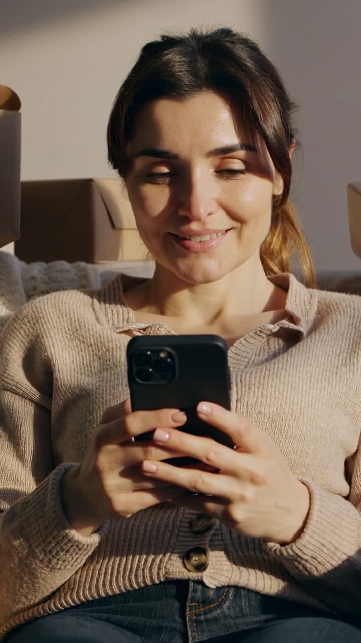Vertical video: Picking up smartphone woman in sweater scrolling and tapping on sofa, with boxes