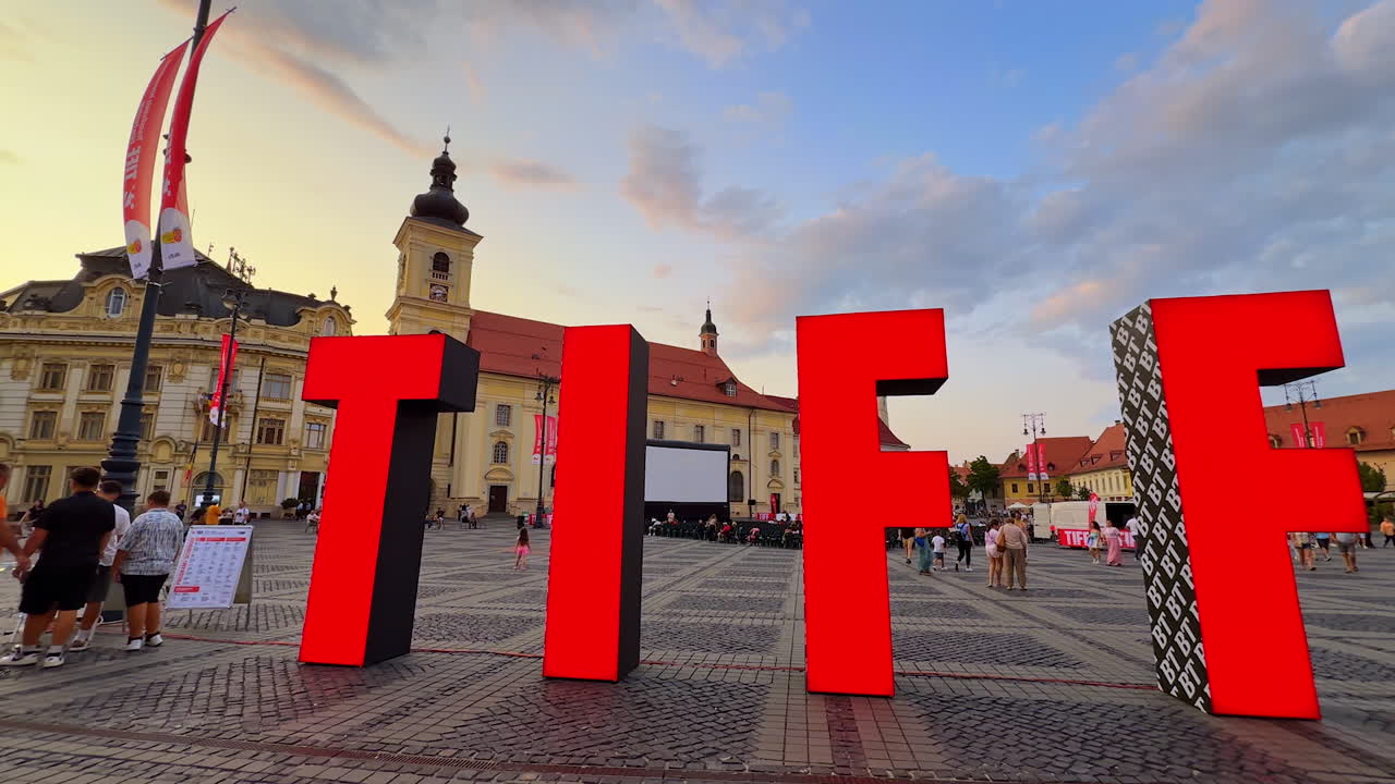 Sibiu, Romania, 17 July 2025: Lively outdoor cinema in Sibiu Main Square at sunset. Church tower and red-tiled roofs frame a large screen with spectators under a dramatic cloudy sky