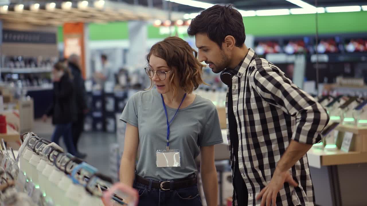 en la tienda de electrónica una consultora da consejos profesionales a un joven. él considera comprar un nuevo teléfono inteligente y necesita la opinión de un experto. la tienda es moderna, brillante y tiene todos los dispositivos nuevos
