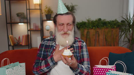 happy elderly old man celebrating birthday party, makes wish blowing burning candle on small cupcake