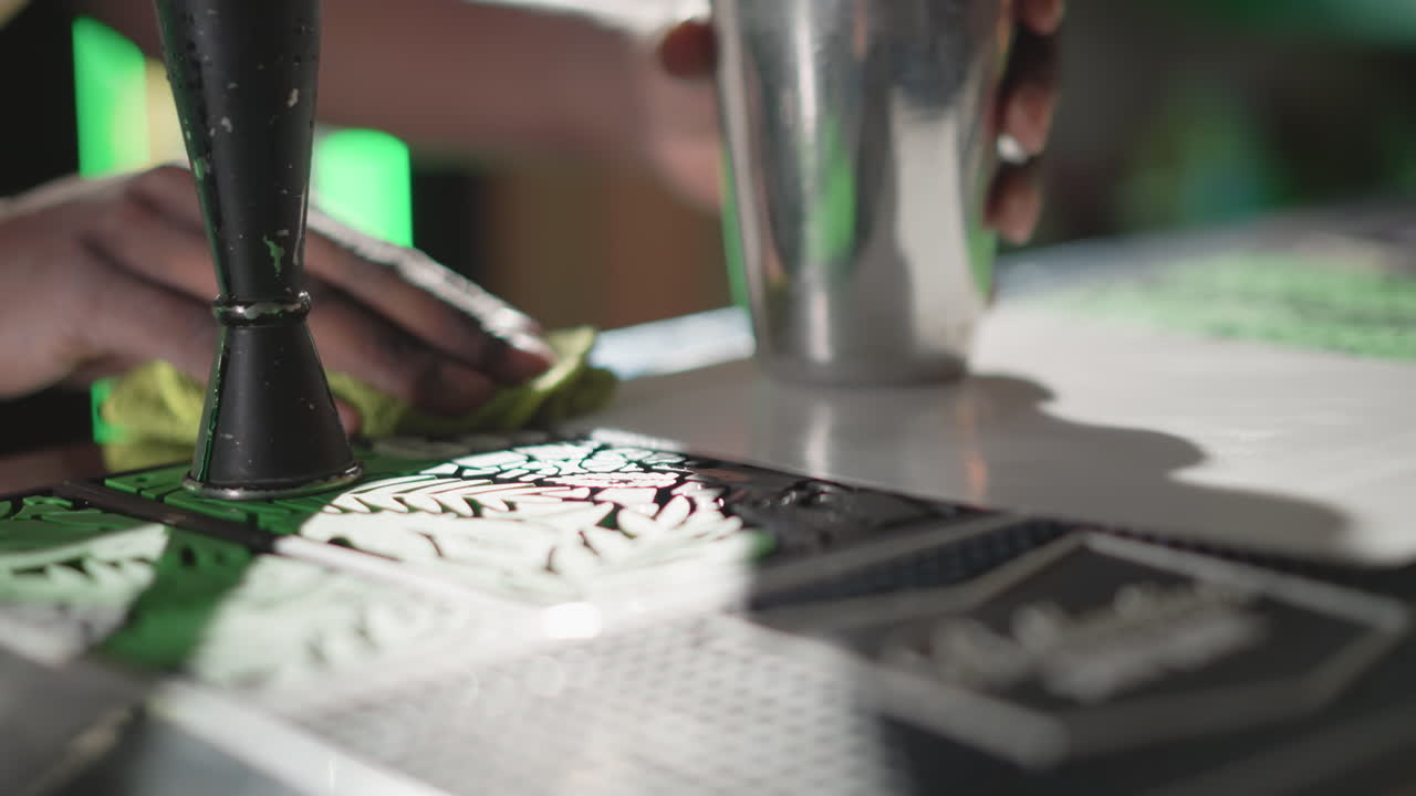 Bartender wiping bar counter with cloth while holding jigger, preparing for drink creation at modern bar with vibrant green lighting, focusing on cleanliness and drink preparation