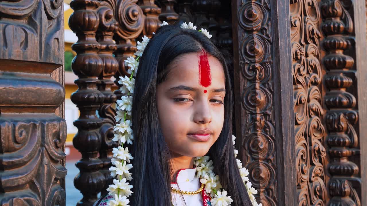 Peaceful young devotee with jasmine flowers in her hair and a red Tikka on her forehead standing near a beautifully carved wooden door of a traditional Nepalese temple