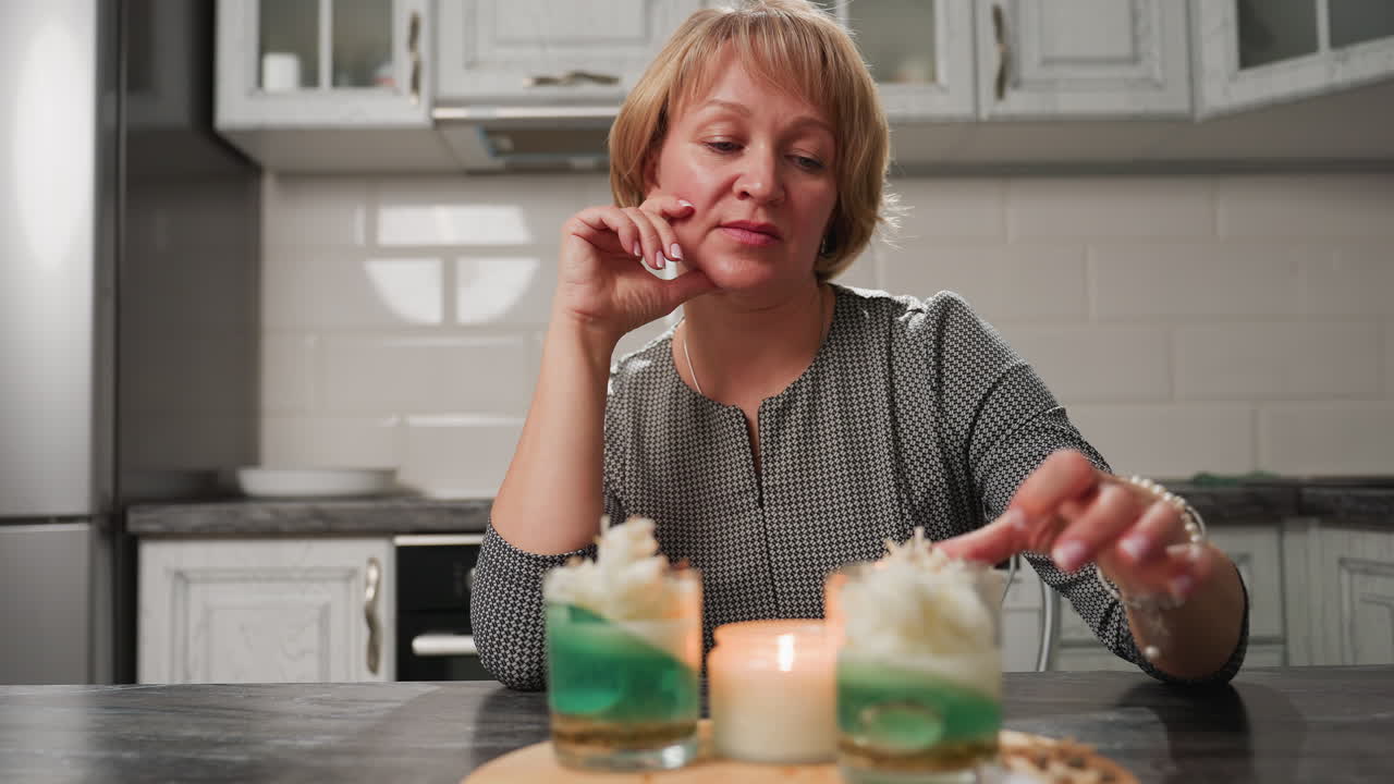 Baker sits at table, one hand wiping cake pan surface, other resting on cheek in thoughtful reflection, focused on baking result. Oven tabletop and rustic kitchen cabinets visible in indoor setting
