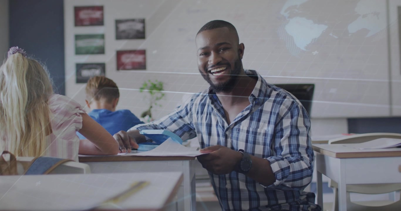 Male teacher kneeling at classroom desk showing paper, blue folder, reviewing student work, smiling