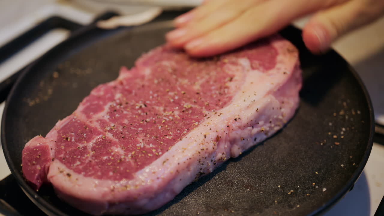 Close up of a raw beef steak being seasoned with salt and black pepper in a pan
