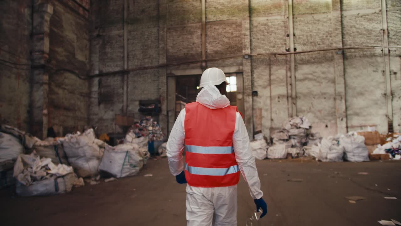 vista trasera de un hombre en un chaleco de seguridad rojo y uniforme blanco caminando a través de una planta de reciclaje de residuos alta. una habitación enorme con osos polares con basura que será reciclada en una planta enorme