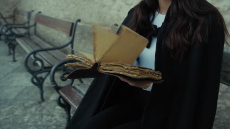 mujer con capa lee los misterios en el libro sentada en un banco en la ciudad