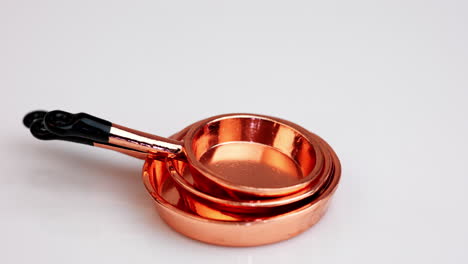 a set of three copper frying pans with black handles piled on each other. miniature crockery at rotating white background.