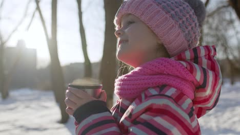 child girl kid drinking hot drink tea from cup on snowy road in winter park, christmas holidays