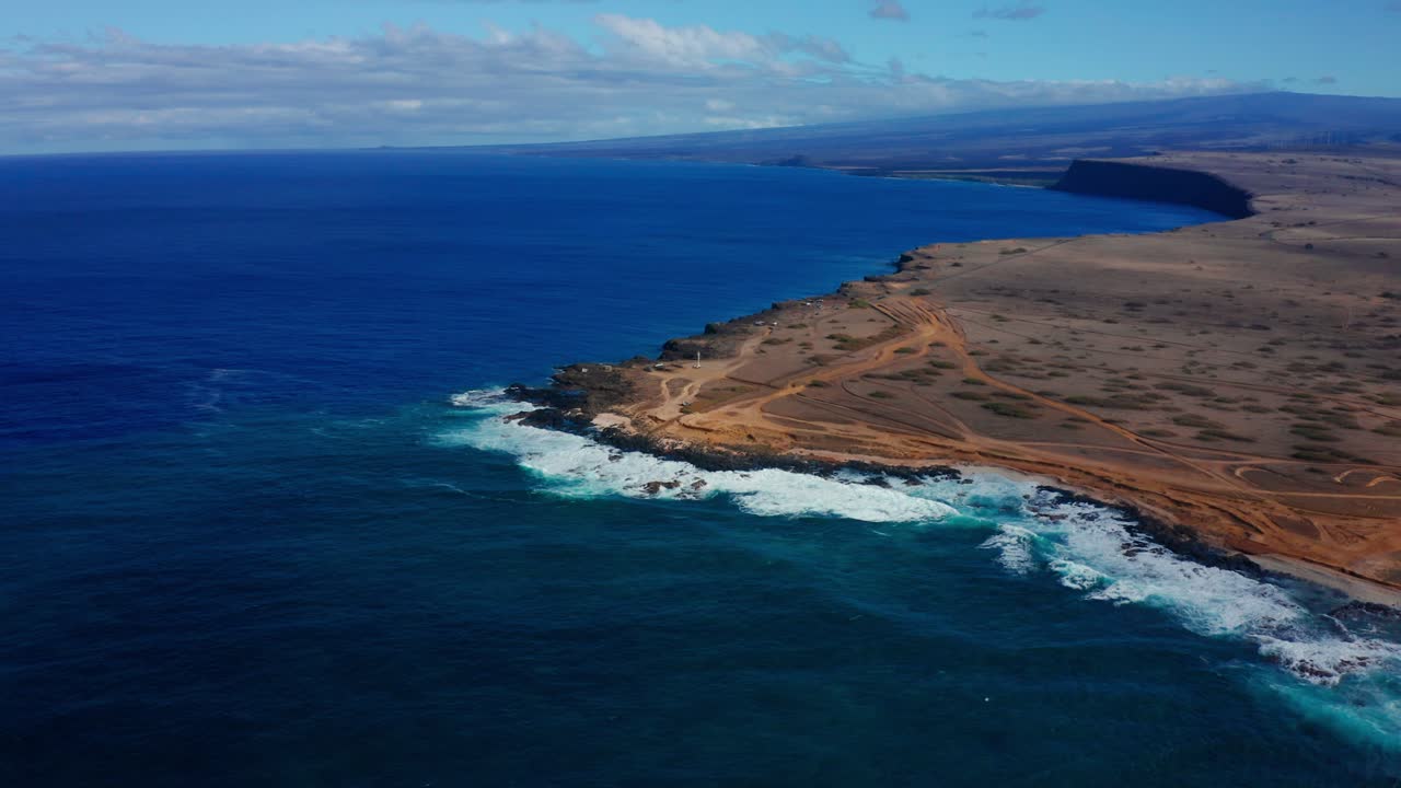 Powerful blue waves batter a barren rocky shoreline traced with dusty paths, creating a raw, windswept coastal landscape under a wide open sky.
