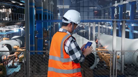 vista trasera de un ingeniero masculino asiático con casco de seguridad trabajando en un teléfono inteligente con el plan de la turbina eólica en una fábrica de turbinas eólicas mientras el brazo robótico trabaja