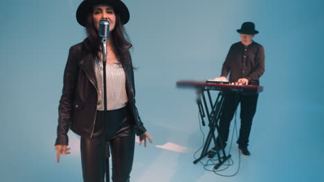 a vocalist in a black leather jacket and hat,smiling and singing with eyes open,stands at a vintage microphone with hands moving up and down a keyboardist works a red sampler against a blue background