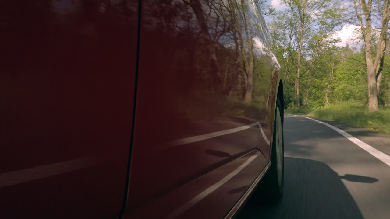 Low side view of red car driving road, reflections of nature on metallic lacquer