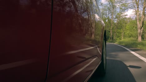 low side view of red car driving road, reflections of nature on metallic lacquer