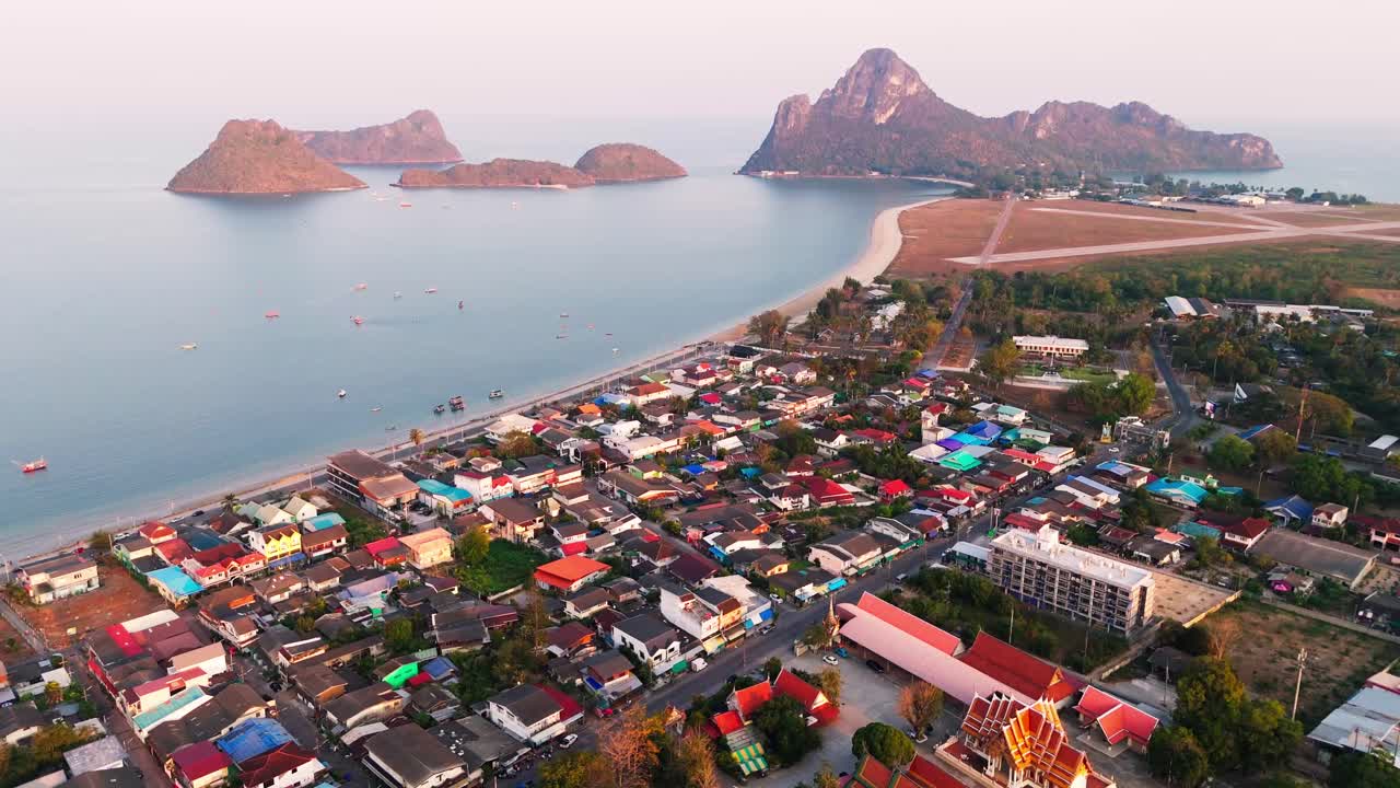 Aerial establishing overview of Prachuap Khiri Khan, the bay bleding with ocean, coastal homes, markets and airstrip near traditional temples as boats anchor in water