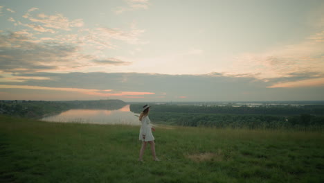 a woman in a white dress and hat walks gracefully through a grassy field towards a serene lake at sunset. she pauses, placing her hands on her ears, creating a tranquil and introspective moment