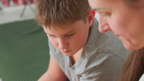 close view of mom and son sitting close together looking serious and focused, woman wearing light beige dress with red earrings, boy wearing grey shirt, indoor setting with green couch background