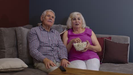 Una-Pareja-Mayor-Disfruta-Viendo-Una-Película-En-La-Televisión-Y-Comiendo-Palomitas-De-Maíz-En-El-Sofá-De-La-Sala-De-Estar-En-Casa.