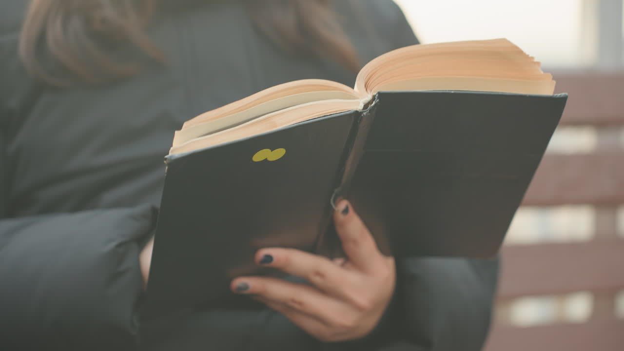 Giovane donna immersa nel suo romanzo preferito. Primo piano delle mani che sfogliano le pagine su una panchina alla luce naturale. Donna con abiti caldi che si gode un momento tranquillo leggendo un tascabile vintage all'aperto.