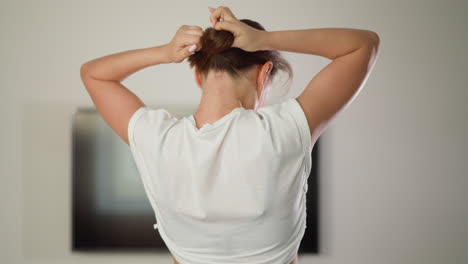 woman ties hair into ponytail in home gym. preparing for jogging. dynamic athlete dressed in sports top arranges short hair running on treadmill indoors