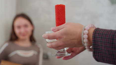 close-up of woman hand holding red candle in glass holder, adorned with elegant beaded bracelet, partial view of lady seated at dining table in cozy, stylish home interior setting