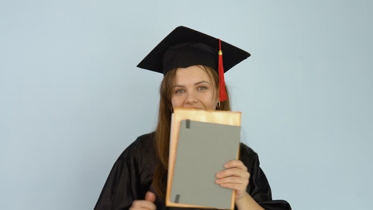 A young female student in a black robe and a master's hat shakes books, which she holds in her hands. White background