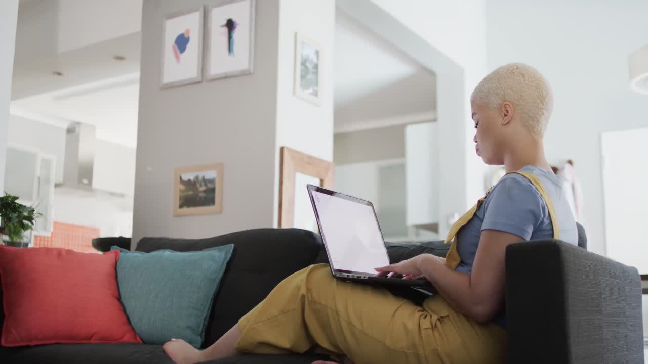 Happy biracial woman using laptop with copy space and sitting on couch in slow motion