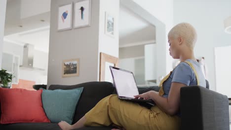 Happy-biracial-woman-using-laptop-with-copy-space-and-sitting-on-couch-in-slow-motion