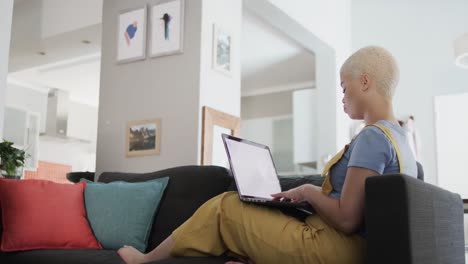mujer biracial feliz usando una computadora portátil con espacio de copia y sentada en el sofá en cámara lenta