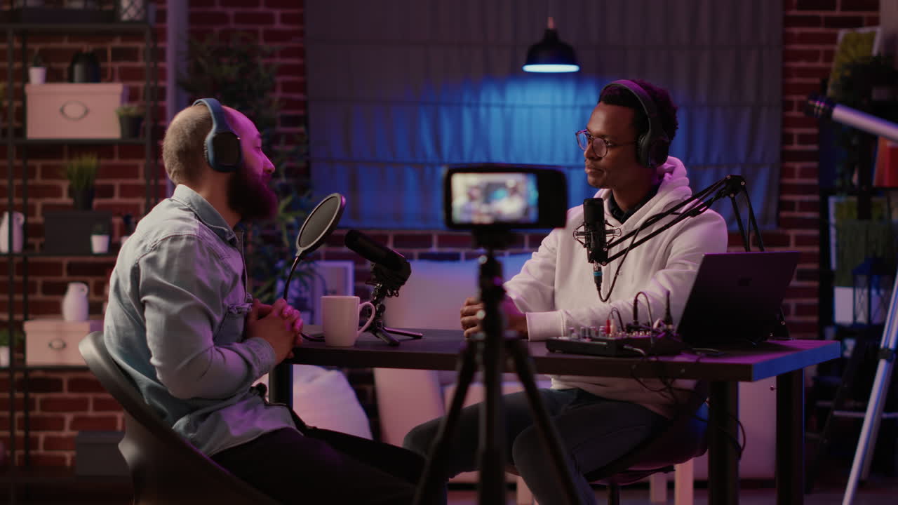 Static tripod shot of internet radio host recording podcast using digital video camera greeting guest