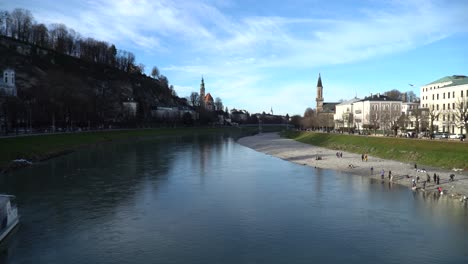 Wide-view-of-Salzach-River-in-Salzburg-with-tourists-on-the-beach-near-the-fortress