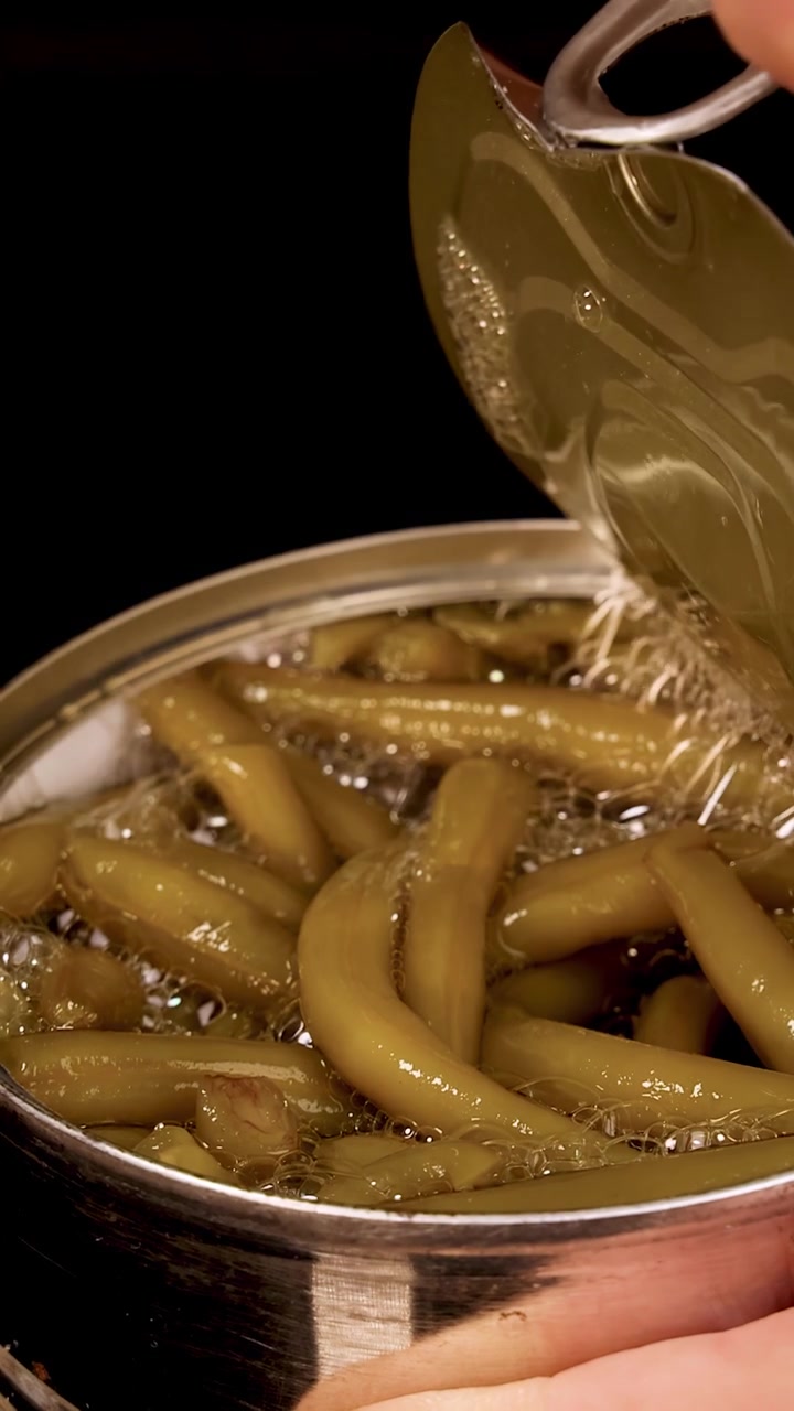 A hand opens a pull-tab can of green beans under bright, even lighting with a close-up perspective and steady camera, revealing the beans inside