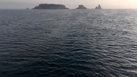 Vista-Aérea-Volando-A-Través-De-Las-Olas-Del-Mar-Mediterráneo-Incline-Hacia-Arriba-Para-Revelar-Las-Islas-Medes-En-El-Horizonte-Del-Amanecer