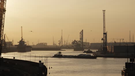 a barge sails through a calm industrial harbor at sunset as birds fly past cranes and silhouetted structures