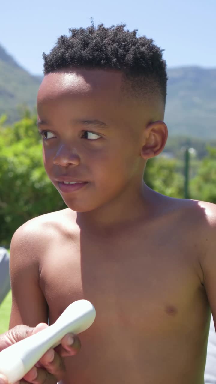 African American dad and son sitting on lounger in yard applying sunscreen in pink trunks smiling
