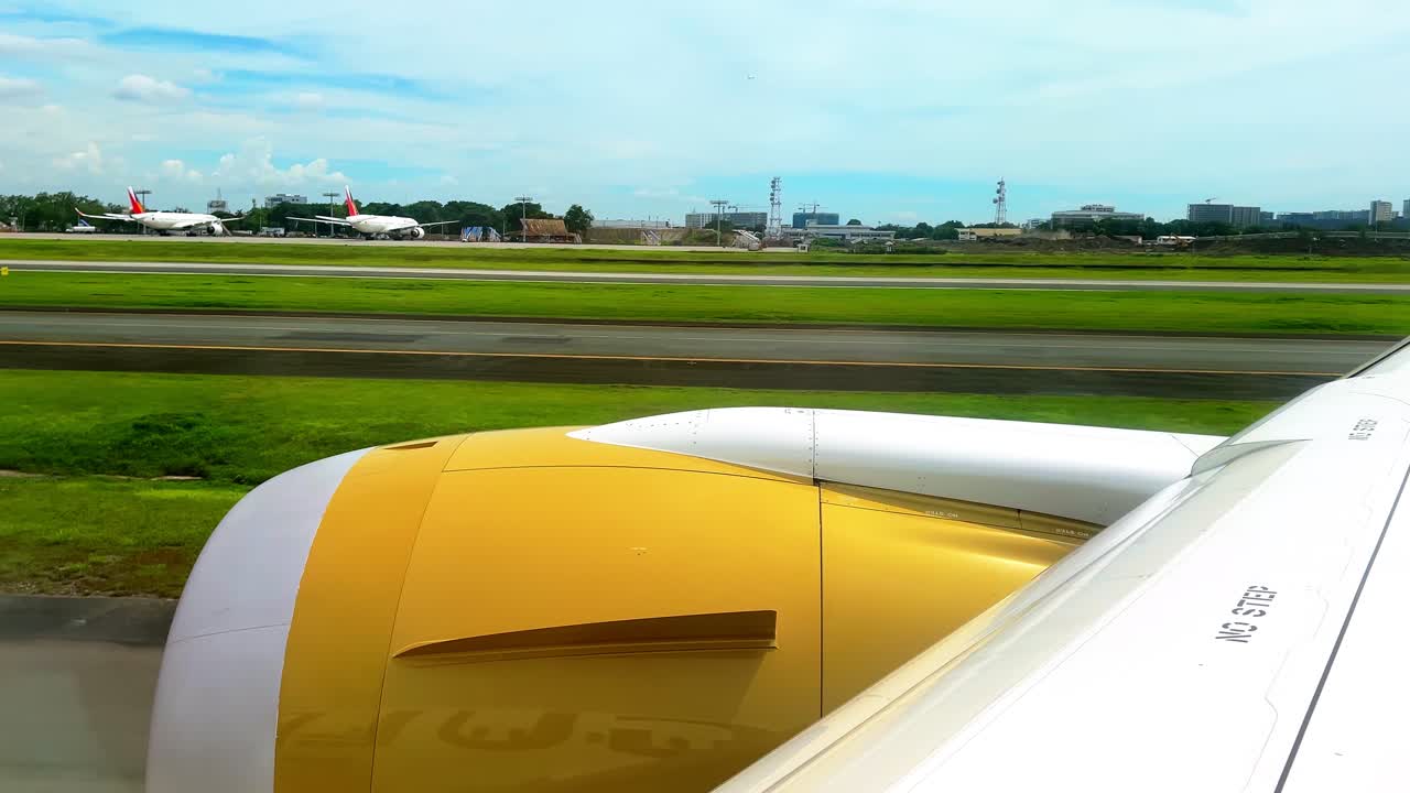 Airplane window view of golden engine and wing lined up on the runway, ready for takeoff, with other aircraft and airport facilities in the distance