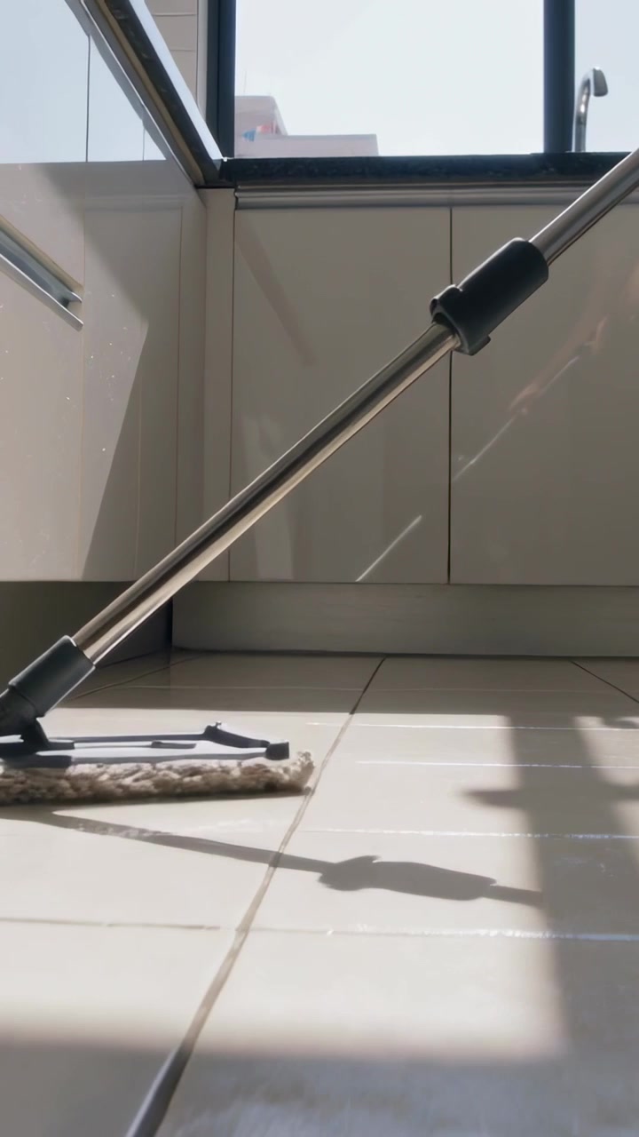 Vertical video: Mopping metal mop sliding over kitchen floor as sun highlighting grout, khaki pants