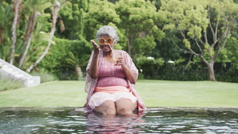 mujer afroamericana senior feliz saludando junto a la piscina con cóctel, espacio de copia, cámara lenta