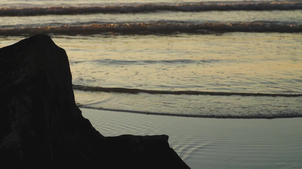 Bølger slår ind mod en sten på stranden under solnedgang.