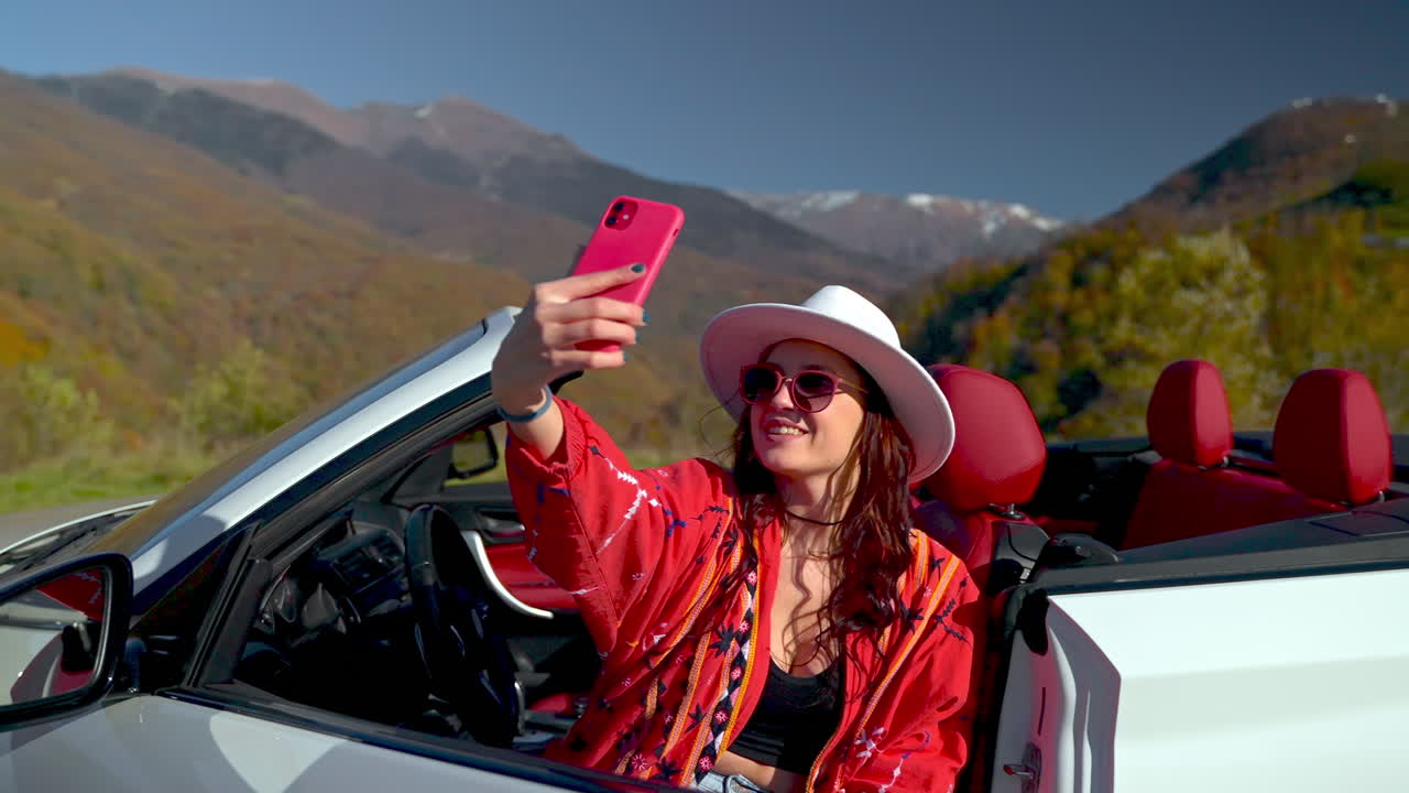 una mujer tomando una selfie en un coche descapotable en una pintoresca carretera de montaña.