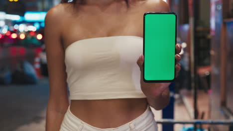 woman holding a smartphone with green screen on a city street at night