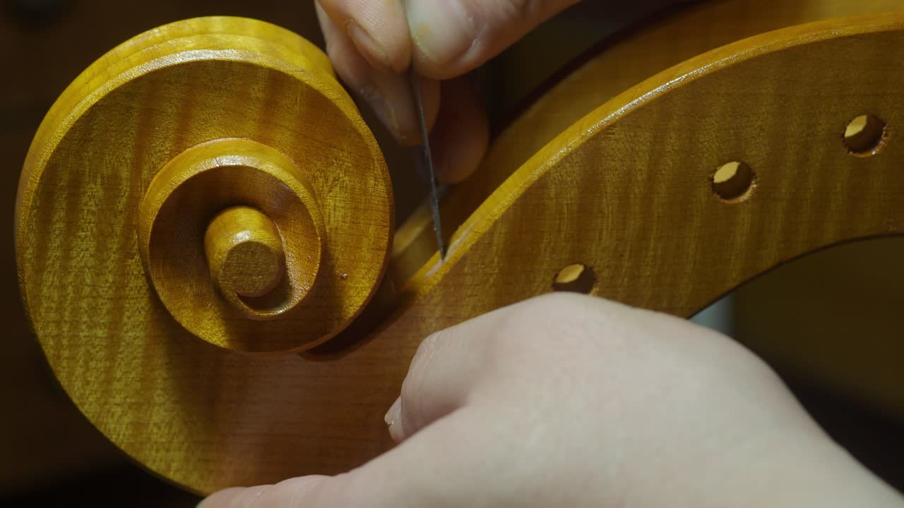 Close-up of a luthier's hands delicately finishing a cello scroll, using a fine tool to carve smooth details into the volute of the instrument during the final stages of craftsmanship